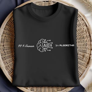 Op de afbeelding: Zwart T-shirt met ronde hals en de tekst "99% Human 1% Algorithm" in het wit. Het ontwerp bevat een hersengrafiek met een AI-symbool. Het shirt is opgevouwen op een rieten mand.