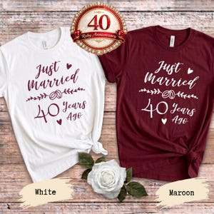 Pode incluir: Uma t-shirt branca com texto bordô e uma t-shirt bordô com texto branco, ambas com a frase Just Married 40 Years Ago, um gráfico de anéis e corações. Um emblema circular no topo diz 40 Ruby Anniversary, com uma rosa branca abaixo.