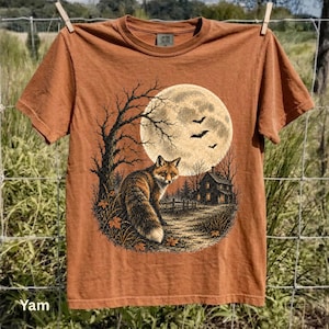 Puede incluir: Una camiseta de manga corta color óxido con un estampado gráfico de un zorro sentado frente a una gran luna llena, un árbol desnudo, una casa pequeña y murciélagos volando. El diseño está en tonos negro, marrón y crema en el centro.