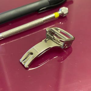 Op de afbeelding: Een roestvrijstalen horlogebandsluiting met een rechthoekige gesp en een gebogen, sleufvormige basis. Een kleine schroevendraaier met een gele punt en een zwarte pen staan ook op de afbeelding, wat wijst op horlogereparatietools.