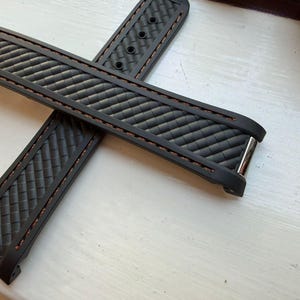 Puede incluir: Una correa de reloj negra con un patr&oacute;n de fibra de carbono texturizado y costuras marrones en los bordes. La correa incluye terminales de metal plateado y una serie de orificios de ajuste circulares para una fijaci&oacute;n segura al reloj.