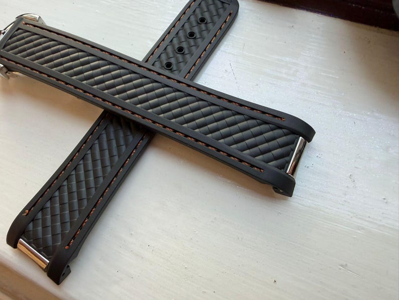 Puede incluir: Una correa de reloj negra con un patr&oacute;n de fibra de carbono texturizado y costuras marrones en los bordes. La correa incluye terminales de metal plateado y una serie de orificios de ajuste circulares para una fijaci&oacute;n segura al reloj.