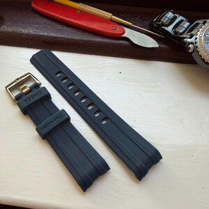 May include: A dark blue rubber watch strap featuring a metal buckle and the embossed text OMEGA and Seamaster on the strap loop and band. The two-piece strap has a smooth finish with vertical ridges and multiple adjustment holes for a secure fit.