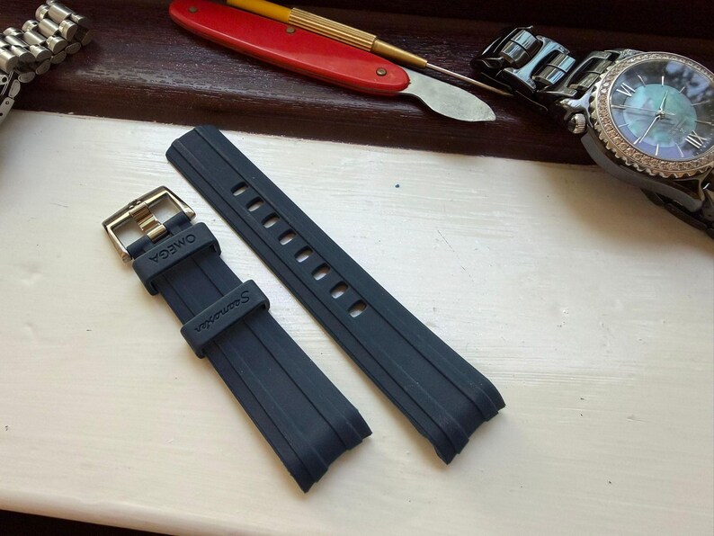 May include: A dark blue rubber watch strap featuring a metal buckle and the embossed text OMEGA and Seamaster on the strap loop and band. The two-piece strap has a smooth finish with vertical ridges and multiple adjustment holes for a secure fit.