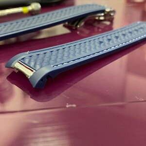 May include: Two blue watch straps with a textured pattern and silver-tone hardware. The straps are on a reflective surface, with a tool in the background. The watch straps are a replacement part.