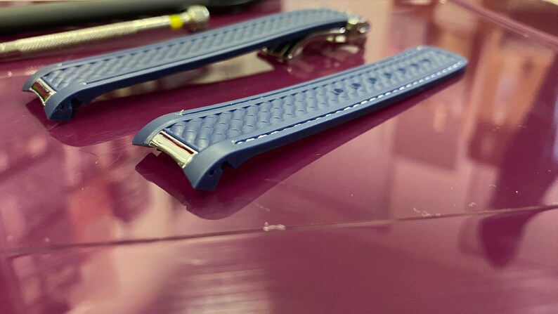 May include: Two blue watch straps with a textured pattern and silver-tone hardware. The straps are on a reflective surface, with a tool in the background. The watch straps are a replacement part.
