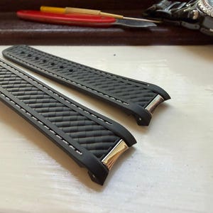 May include: A pair of black rubber watch straps featuring a textured basketweave pattern on the surface, white contrast stitching along the edges, and polished silver-tone metal end links designed for attachment to a watch case.