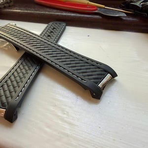 May include: A black rubber watch strap featuring a textured carbon fibre pattern, white contrast stitching along the edges, and polished silver-tone metal end links designed to fit flush against a watch case. The strap has a flexible construction.