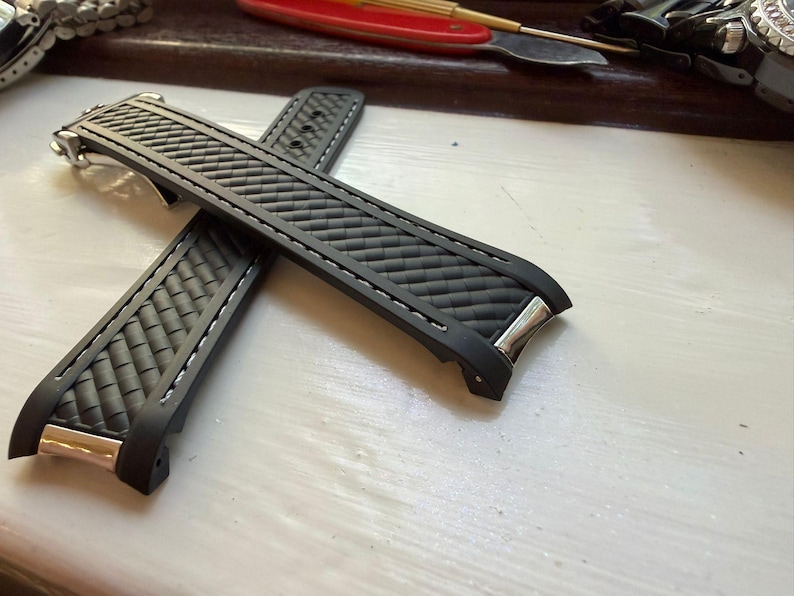 May include: A black rubber watch strap featuring a textured carbon fibre pattern, white contrast stitching along the edges, and polished silver-tone metal end links designed to fit flush against a watch case. The strap has a flexible construction.