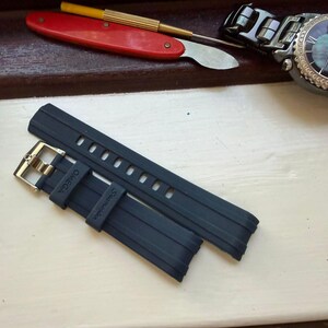 May include: A dark blue rubber watch strap featuring a silver-toned metal buckle. The strap is embossed with the word OMEGA and the word Seamaster on the keeper. The design includes a series of rectangular adjustment holes for a secure fit.