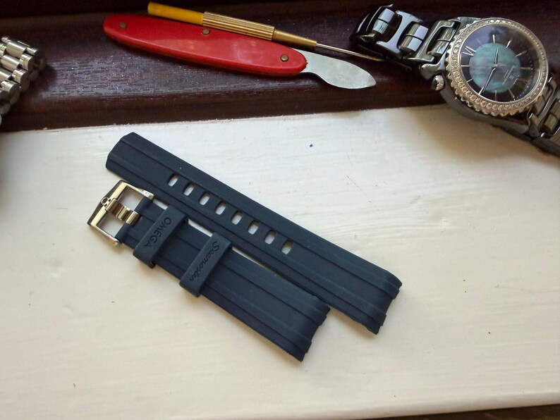 May include: A dark blue rubber watch strap featuring a silver-toned metal buckle. The strap is embossed with the word OMEGA and the word Seamaster on the keeper. The design includes a series of rectangular adjustment holes for a secure fit.