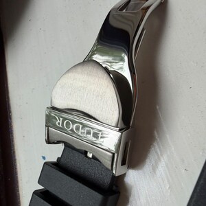K&ouml;nnte beinhalten: Eine silberfarbene Uhren-Faltschlie&szlig;e aus Edelstahl mit geb&uuml;rstetem und poliertem Finish, auf deren Vorderseite das Wort TUDOR eingraviert ist. Die Schlie&szlig;e ist an einem schwarzen Kautschuk-Uhrenarmband mit zwei integrierten Kautschukschlaufen befestigt.