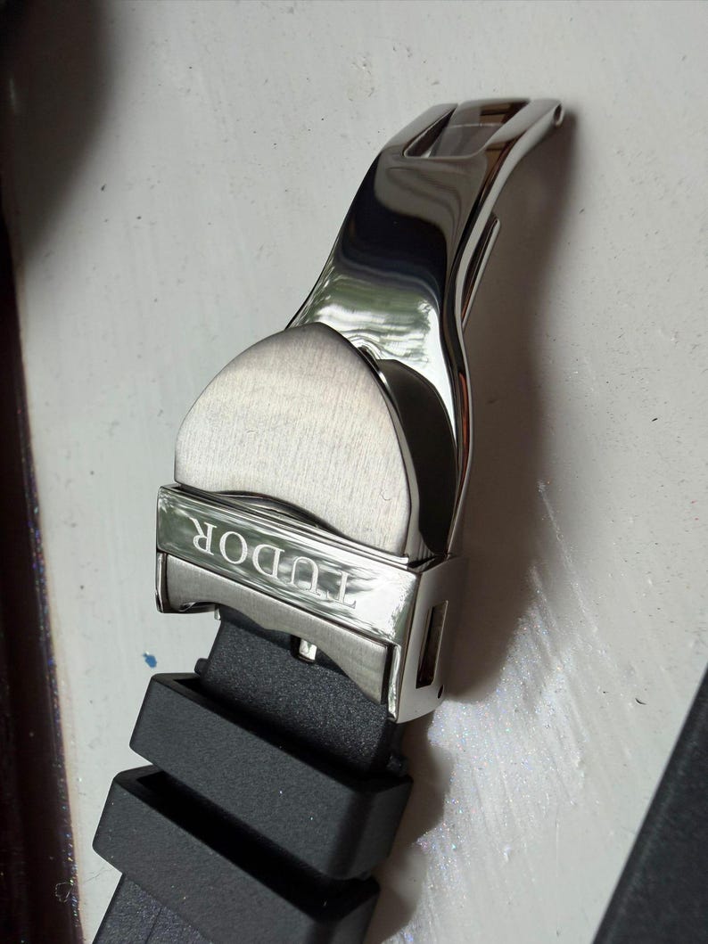 K&ouml;nnte beinhalten: Eine silberfarbene Uhren-Faltschlie&szlig;e aus Edelstahl mit geb&uuml;rstetem und poliertem Finish, auf deren Vorderseite das Wort TUDOR eingraviert ist. Die Schlie&szlig;e ist an einem schwarzen Kautschuk-Uhrenarmband mit zwei integrierten Kautschukschlaufen befestigt.