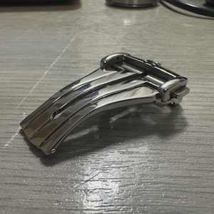 May include: A polished silver-tone stainless steel watch deployment clasp featuring a curved, multi-link design with a secure folding mechanism. The metal surface is highly reflective, showcasing a smooth, contoured shape with visible hinge points.