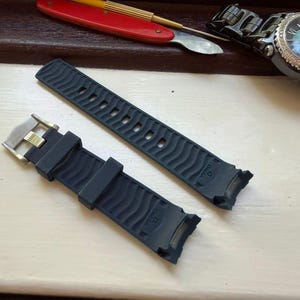 May include: A dark blue rubber watch strap featuring a wavy textured pattern and the Omega brand name embossed on the surface. The strap includes a silver-toned metal buckle and curved ends designed for attachment to a watch case.