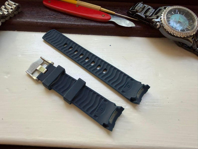 May include: A dark blue rubber watch strap featuring a wavy textured pattern and the Omega brand name embossed on the surface. The strap includes a silver-toned metal buckle and curved ends designed for attachment to a watch case.