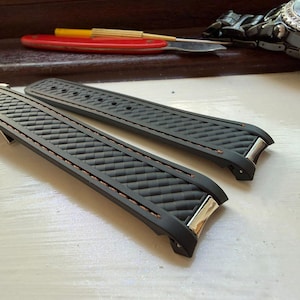 Puede incluir: Un par de correas de reloj de caucho negro con un patr&oacute;n texturizado de fibra de carbono en la superficie, acentuadas con costuras finas de color naranja a lo largo de los bordes y terminales de metal plateado para el reloj.