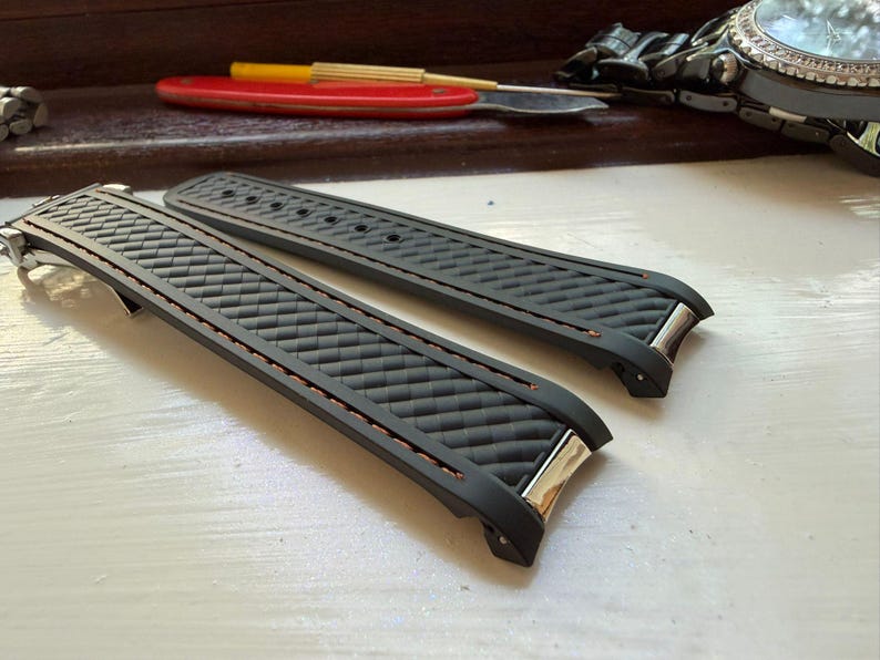 Puede incluir: Un par de correas de reloj de caucho negro con un patr&oacute;n texturizado de fibra de carbono en la superficie, acentuadas con costuras finas de color naranja a lo largo de los bordes y terminales de metal plateado para el reloj.