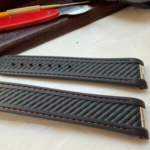 Puede incluir: Una correa de reloj negra con un patr&oacute;n de textura diagonal y costuras marrones a lo largo de los bordes. La correa consta de dos piezas separadas, cada una con accesorios de metal plateado en los extremos y varios orificios de ajuste.