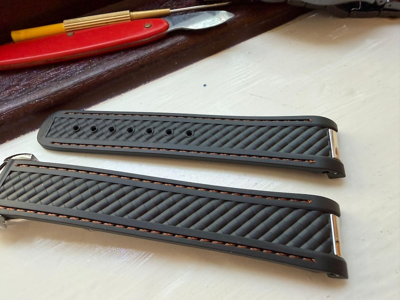 Puede incluir: Una correa de reloj negra con un patr&oacute;n de textura diagonal y costuras marrones a lo largo de los bordes. La correa consta de dos piezas separadas, cada una con accesorios de metal plateado en los extremos y varios orificios de ajuste.