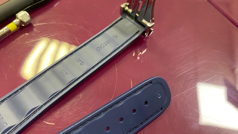 May include: A close-up of a navy blue watch strap with black stitching and a metal clasp. The strap has the number "20" printed on it, along with the text "GJD" and "CV200179".