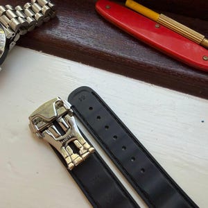 May include: A black rubber watch strap featuring a silver-toned metal deployment clasp with the engraved number 04521819. The strap includes a series of adjustment holes and is positioned near a wristwatch and a red-handled tool.
