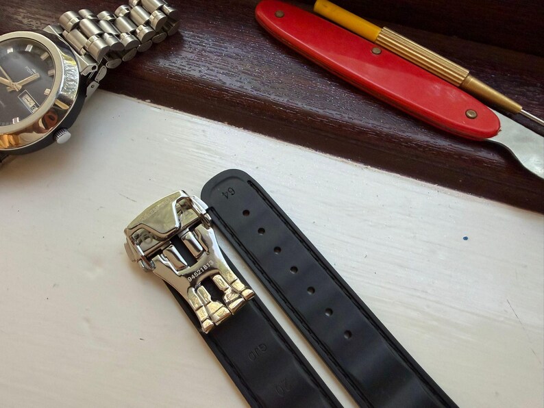 May include: A black rubber watch strap featuring a silver-toned metal deployment clasp with the engraved number 04521819. The strap includes a series of adjustment holes and is positioned near a wristwatch and a red-handled tool.