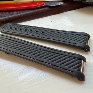 Puede incluir: Una correa de reloj negra con un patr&oacute;n diagonal texturizado y costuras marrones en los bordes. La correa incluye herrajes met&aacute;licos de color plateado en los puntos de fijaci&oacute;n y una serie de orificios circulares para ajustar en la pieza larga.