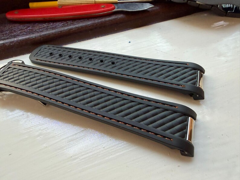 Puede incluir: Una correa de reloj negra con un patr&oacute;n diagonal texturizado y costuras marrones en los bordes. La correa incluye herrajes met&aacute;licos de color plateado en los puntos de fijaci&oacute;n y una serie de orificios circulares para ajustar en la pieza larga.