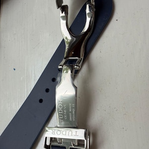 K&ouml;nnte beinhalten: Ein dunkelblaues Kautschuk-Uhrenarmband mit einer silberfarbenen Edelstahl-Faltschlie&szlig;e. Die Metallschlie&szlig;e ist mit dem Text TUDOR, GENEVE, SWISS MADE und STEELINOX EB graviert. Das Armband verf&uuml;gt &uuml;ber eine Reihe von runden Einstelll&ouml;chern.
