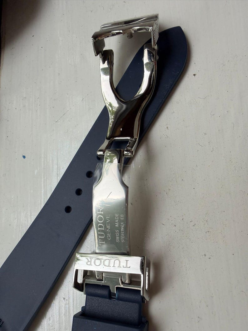 K&ouml;nnte beinhalten: Ein dunkelblaues Kautschuk-Uhrenarmband mit einer silberfarbenen Edelstahl-Faltschlie&szlig;e. Die Metallschlie&szlig;e ist mit dem Text TUDOR, GENEVE, SWISS MADE und STEELINOX EB graviert. Das Armband verf&uuml;gt &uuml;ber eine Reihe von runden Einstelll&ouml;chern.