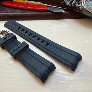 May include: A black rubber watch strap featuring a silver-toned metal buckle. The strap is embossed with the text OMEGA on the keeper and Seamaster on the band. The design includes a series of rectangular adjustment holes and raised parallel lines.
