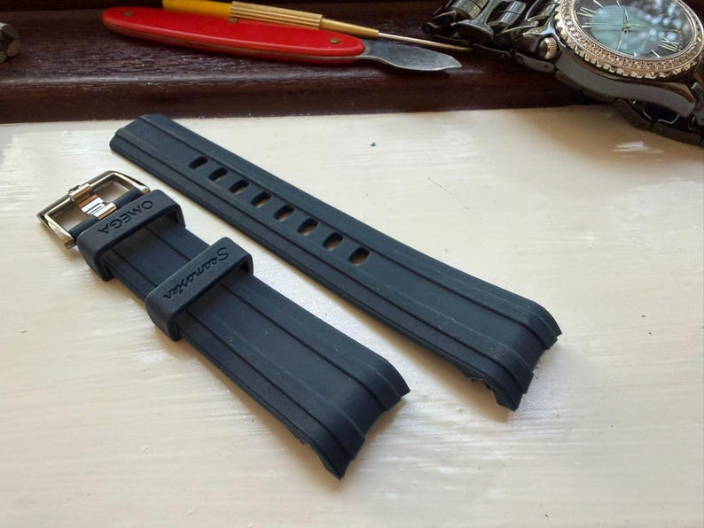May include: A black rubber watch strap featuring a silver-toned metal buckle. The strap is embossed with the text OMEGA on the keeper and Seamaster on the band. The design includes a series of rectangular adjustment holes and raised parallel lines.