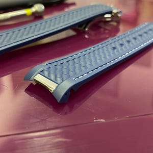 May include: Two blue watch straps with a textured pattern and white stitching. The straps have silver-coloured metal accents. They are on a purple surface. A small tool is visible in the background.