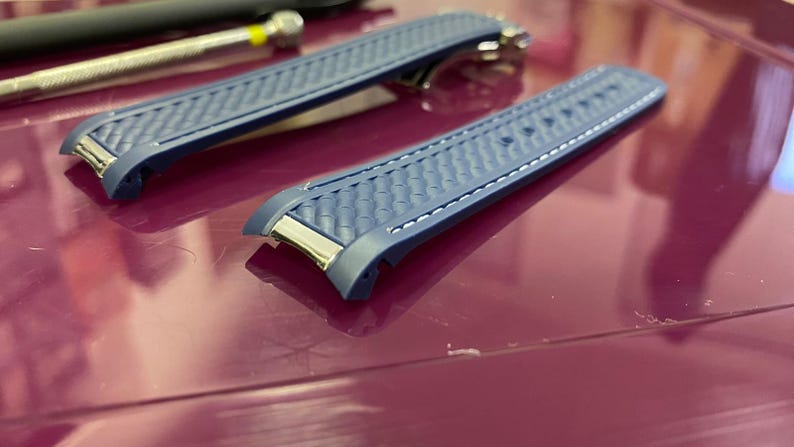 May include: Two blue watch straps with a textured pattern and white stitching. The straps have silver-coloured metal accents. They are on a purple surface. A small tool is visible in the background.