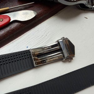 May include: A black rubber watch strap featuring a silver-toned stainless steel deployment clasp with the text TAGHeuer, FC 5030, and STAINLESS STEEL F 50 engraved on the metal components. The strap has a perforated texture on one side.