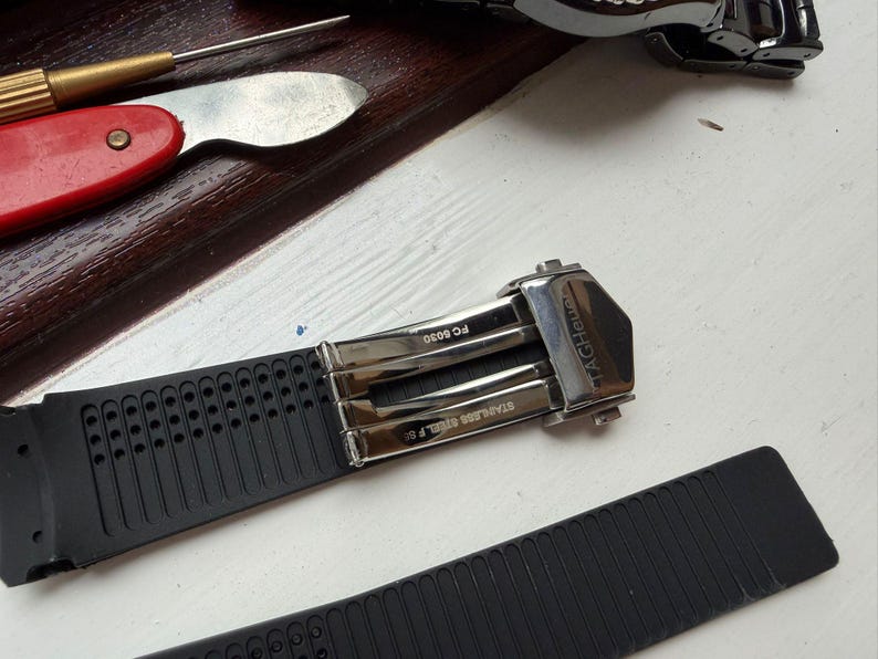 May include: A black rubber watch strap featuring a silver-toned stainless steel deployment clasp with the text TAGHeuer, FC 5030, and STAINLESS STEEL F 50 engraved on the metal components. The strap has a perforated texture on one side.