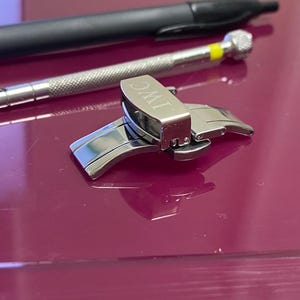 May include: A silver watch clasp with the engraved letters "IWC" is the focal point of this image. A small screwdriver and a pen are also present. The objects are on a reflective, maroon surface.