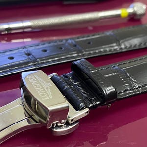 May include: A close-up of a black leather watch strap with a silver clasp, bearing the "Longines" brand name. A watch repair tool is in the background. The strap has a textured pattern and is designed for a wristwatch.