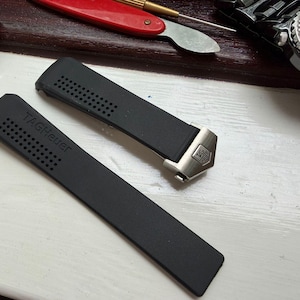 May include: A black rubber watch strap featuring a perforated texture and the embossed text TAGHeuer. The strap includes a silver-toned metal end link with an engraved TAGHeuer logo, designed for attachment to a watch case.
