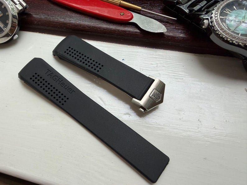May include: A black rubber watch strap featuring a perforated texture and the embossed text TAGHeuer. The strap includes a silver-toned metal end link with an engraved TAGHeuer logo, designed for attachment to a watch case.