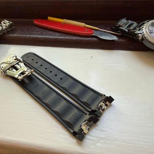May include: A black rubber watch strap featuring a silver-toned metal deployment clasp. The strap is embossed with the Omega logo and the number 20. The design includes a series of horizontal ridges along the length of the band.
