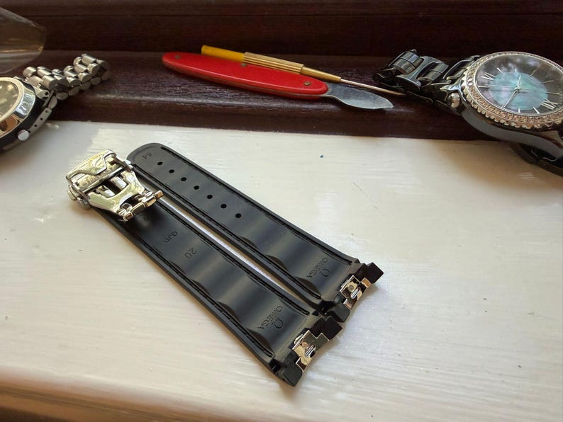 May include: A black rubber watch strap featuring a silver-toned metal deployment clasp. The strap is embossed with the Omega logo and the number 20. The design includes a series of horizontal ridges along the length of the band.