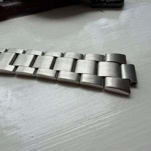 May include: A silver-toned stainless steel watch band featuring a brushed finish with rectangular interlocking links. The metal bracelet includes a folding clasp mechanism at one end and a series of uniform, flat-surfaced links that connect to form a flexible strap for a wristwatch.