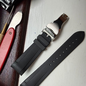 K&ouml;nnte beinhalten: Ein schwarzes Kautschuk-Uhrenarmband mit einer silberfarbenen Metall-Faltschlie&szlig;e, in die das Wort TUDOR eingraviert ist. Das Armband besteht aus zwei separaten Teilen, einem mit dem Schlie&szlig;mechanismus und einem mit einer Reihe von Einstelll&ouml;chern.