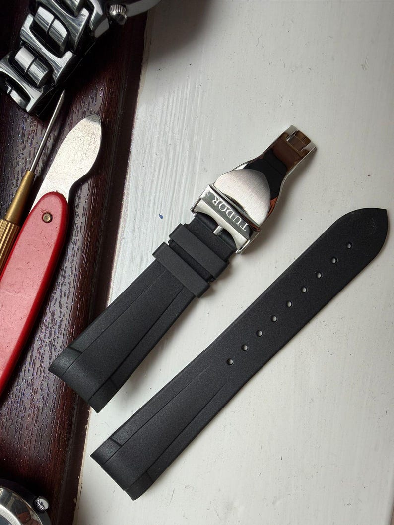 K&ouml;nnte beinhalten: Ein schwarzes Kautschuk-Uhrenarmband mit einer silberfarbenen Metall-Faltschlie&szlig;e, in die das Wort TUDOR eingraviert ist. Das Armband besteht aus zwei separaten Teilen, einem mit dem Schlie&szlig;mechanismus und einem mit einer Reihe von Einstelll&ouml;chern.