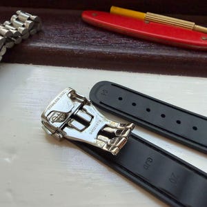 Puede incluir: Una correa de reloj de caucho negro con un cierre desplegable de acero inoxidable plateado. El cierre tiene grabado el texto STAINLESS STEEL y el n&uacute;mero 94521815. La correa incluye las marcas GJD y 20 grabadas en la superficie interior.