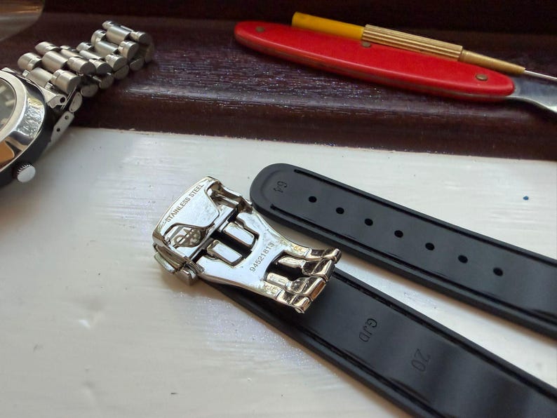Puede incluir: Una correa de reloj de caucho negro con un cierre desplegable de acero inoxidable plateado. El cierre tiene grabado el texto STAINLESS STEEL y el n&uacute;mero 94521815. La correa incluye las marcas GJD y 20 grabadas en la superficie interior.