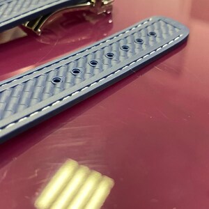 May include: Close-up of a blue watch strap with a textured pattern and white stitching. The strap has several holes for adjustment and a metal clasp is visible in the background. The strap is lying on a red surface.
