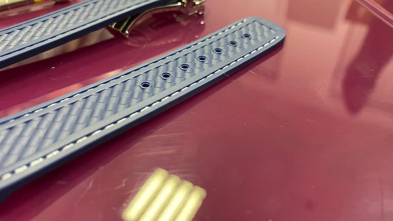 May include: Close-up of a blue watch strap with a textured pattern and white stitching. The strap has several holes for adjustment and a metal clasp is visible in the background. The strap is lying on a red surface.